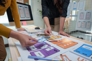 Two female ux designers are collaborating on a project, arranging wireframes on a table to design the user interface and experience for a mobile application